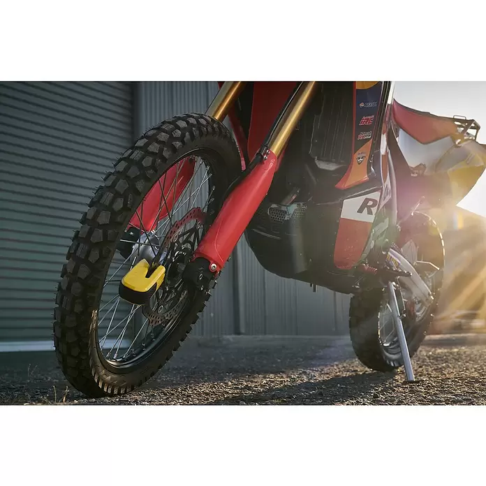 Abus Bremsscheibenschloss GRANIT™ Detecto One 8078 2.0 - Geeignet für: Motorrad, Gelb - Nahaufnahme eines rot-goldenen Rallye-Motorrads mit Stollenreifen, Scheibenbremse und gelbem Bremsscheibenschloss, in der Sonne geparkt.