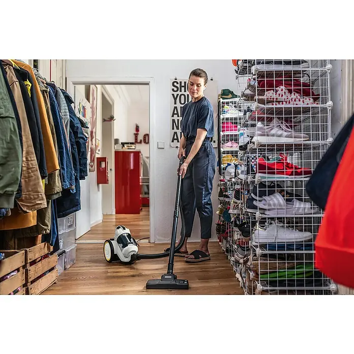 Kärcher Elektro-Staubsauger VC 3 - 700 W - Eine Frau staubsaugt mit einem Kärcher Staubsauger einen Holzboden in einem Flur mit Schuhregalen und einer Garderobe.