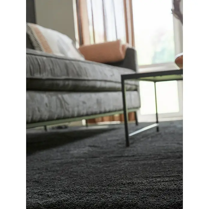 Teppichfliese Amazing - Hochflor, 100 % Polyester, Anthrazit - Nahaufnahme eines strukturierten, dunklen Teppichs im Wohnzimmer, neben einem Sofa und einem Tisch, der Gemütlichkeit und Komfort ausstrahlt.