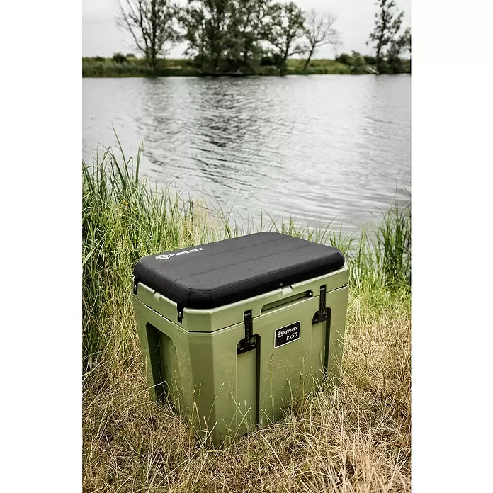 Petromax Nevera de camping - Verde oliva, 25 l, L x An x Al: 58 x 40,5 x 45 cm - Nevera Petromax kx50 verde oliva con cojín negro, junto a un lago entre hierba alta.