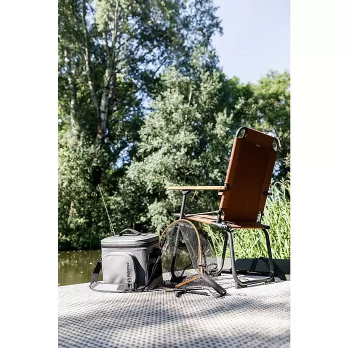 Petromax Nevera portátil - Gris oscuro, 22 l, L x An x Al: 42,5 x 34 x 35,5 cm - Nevera plegable gris, silla de pesca marrón y red de pesca junto a un lago.
