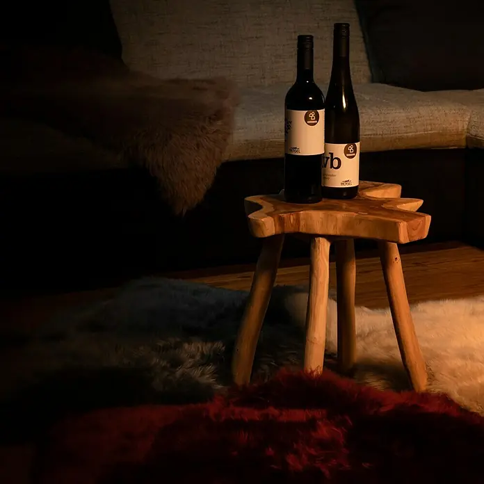 Lammfell - Altrosa, Länge: 90 cm, 100 % Echtfell - Zwei Flaschen Hensel Wein auf einem Holzhocker, umgeben von Teppichen. Perfekt für einen gemütlichen Abend.
