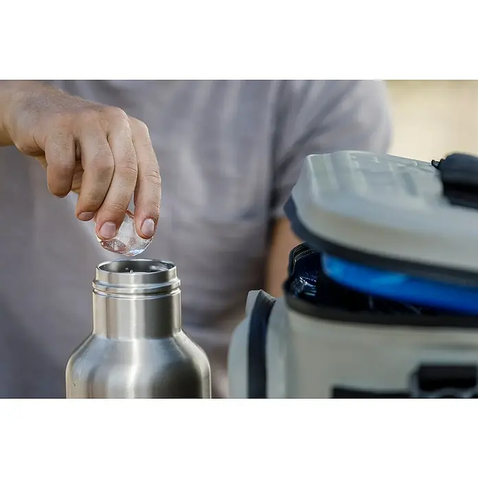 Petromax Thermoflasche - 1 l, Edelstahl, Ø x H: 11,2 x 25,1 cm - Eine Person fügt Eiswürfel in eine Edelstahl-Wasserflasche. Daneben steht eine Kühlbox für unterwegs, um Getränke kühl zu halten.