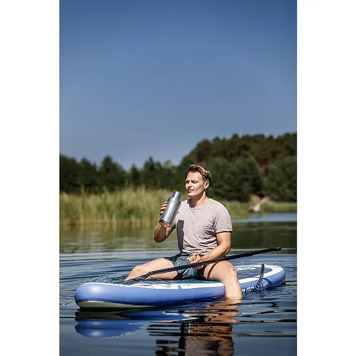 Petromax Thermoflasche - 1,5 l, Edelstahl, Ø x H: 11,2 x 32,2 cm - Ein Mann sitzt auf einem Stand-Up-Paddleboard und trinkt aus einer Wasserflasche. Das Board ist blau und weiß.
