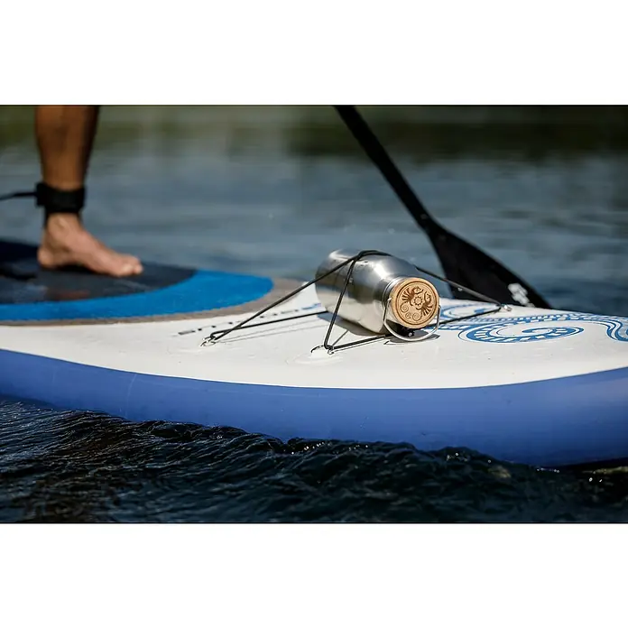 Petromax Thermoflasche - 1,5 l, Edelstahl, Ø x H: 11,2 x 32,2 cm - Eine wiederverwendbare Edelstahlflasche mit Holzdeckel liegt auf einem Paddleboard, befestigt mit einer schwarzen Kordel. Wassersport mit Stil.