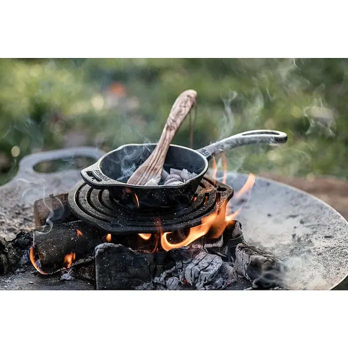 Petromax Gussrost Einsatz - Durchmesser: 23 cm, Gusseisen - Eine kleine gusseiserne Pfanne mit Holzlöffel steht auf einer Feuerstelle mit brennendem Holz, ideal für Outdoor-Kochabenteuer.