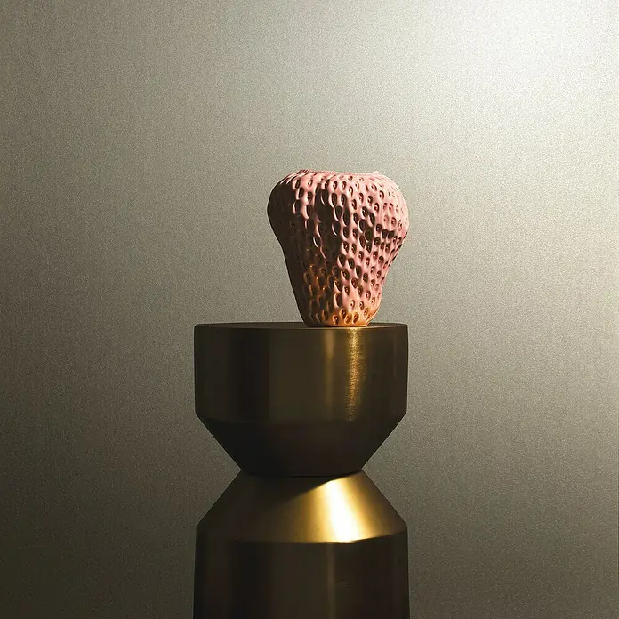 Marburg Eclectic Vliestapete - Graubeige, Metalloptik, 10,05 x 0,7 m - Eine Nahaufnahme einer skulpturalen Erdbeer-Vase in Rosa, die auf einem schlanken, goldenen Sockel steht, vor einem neutralen Hintergrund.