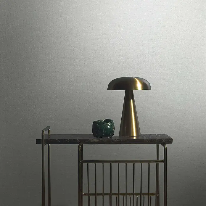 Marburg Eclectic Vliestapete - Helles Beige, Uni, 10,05 x 0,7 m - Nahaufnahme einer Tischlampe und einer dekorativen Dose auf einem goldenen Beistelltisch vor einer grauen Wand.