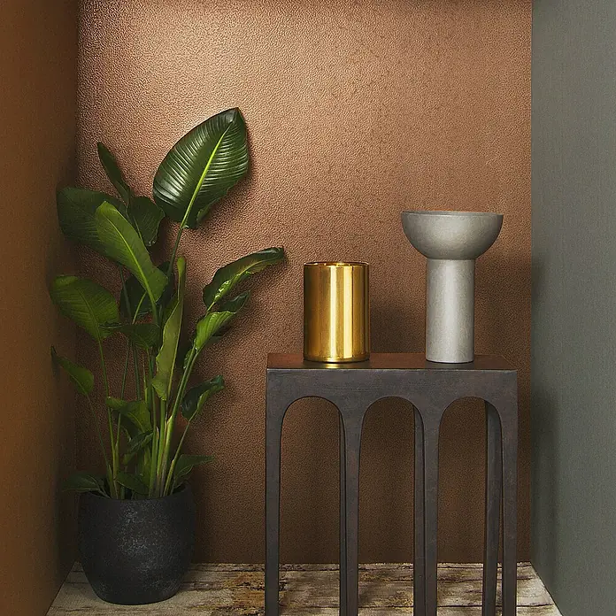 Marburg Eclectic Vliestapete Hammerschlag-Optik - Kupfer, Metalloptik, 10,05 x 0,7 m - Einrichtungsszene mit einer Grünpflanze, einem goldenen Zylinder und einer grauen Schale auf einem dunklen Beistelltisch vor einer braunen Wand.