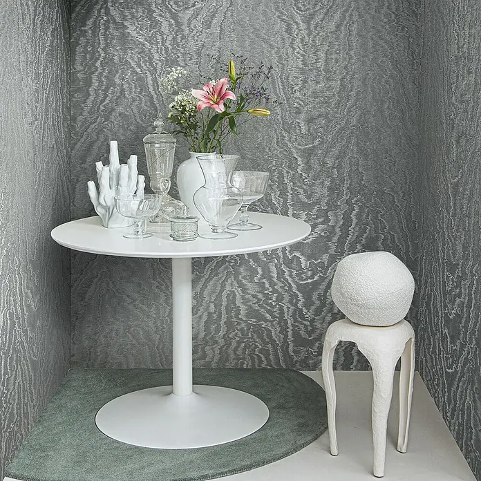 Marburg Eclectic Vliestapete Moiré - Anthrazit, Metalloptik, 10,05 x 0,7 m - Ein weißer runder Tisch mit Blumenvase, Gläsern und Deko-Elementen steht auf einem grünen Teppich. Ein Hocker mit weißer Kugel steht daneben.