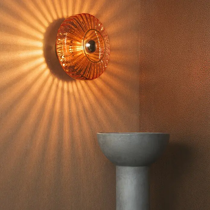Marburg Eclectic Vliestapete Hammerschlag-Optik - Kupfer, Metalloptik, 10,05 x 0,7 m - Eine bernsteinfarbene Wandleuchte wirft ein sternförmiges Lichtmuster auf die Wand und erhellt einen grauen Sockel.