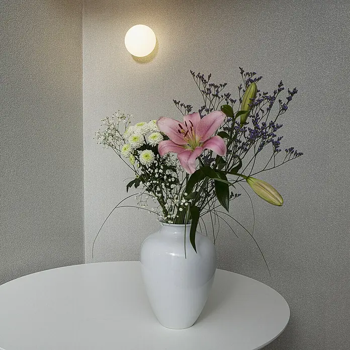 Marburg Eclectic Vliestapete - Sandbeige, Uni, 10,05 x 0,7 m - Nahaufnahme einer weißen Vase mit einem Blumenarrangement auf einem weißen Tisch vor einer grauen Wand mit einer runden Lampe.
