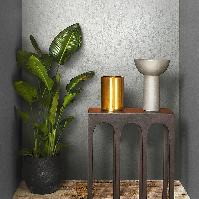 Marburg Eclectic Vliestapete Glasperlenstruktur - Lichtgrau, Metalloptik, 10,05 x 0,7 m - Elegantes Arrangement mit einer goldenen Vase und einem stilvollen Gefäß auf einem dunklen Beistelltisch neben einer üppigen Zimmerpflanze.