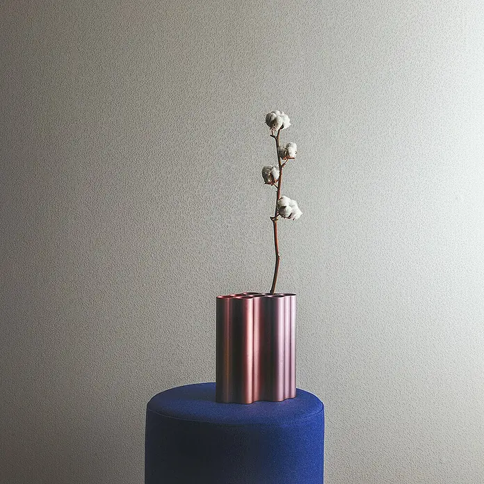 Marburg Eclectic Vliestapete Hammerschlag-Optik - Weiß, Metalloptik, 10,05 x 0,7 m - Eine minimalistische Szene mit einer dekorativen Vase mit Baumwollzweig, die auf einem blauen Hocker steht, vor einem hellen Hintergrund.