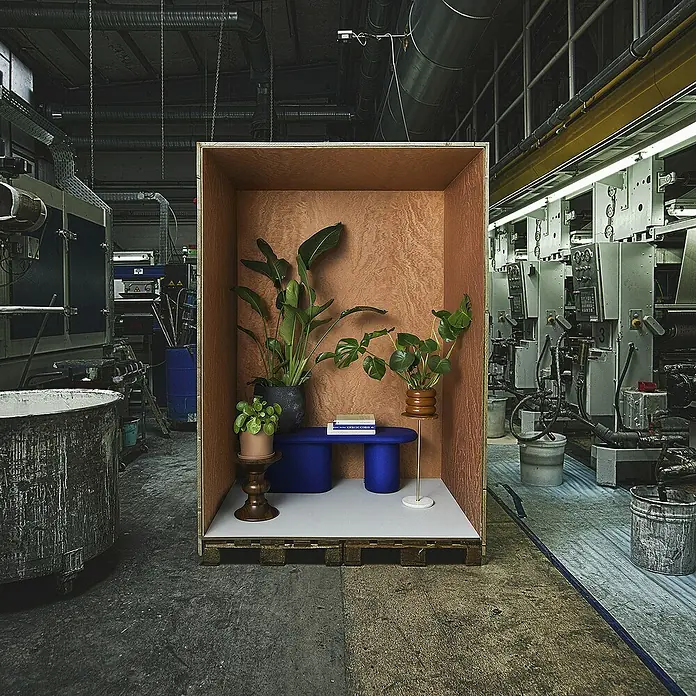 Marburg Eclectic Vliestapete Moiré - Kupfer, Metalloptik, 10,05 x 0,7 m - Zimmer im Zimmer: Ein Würfel mit Pflanzen, Bücher und einer blauen Bank steht in einer Industriehalle.