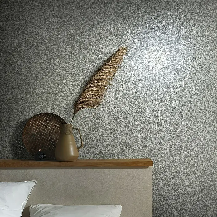 Marburg Eclectic Vliestapete Hammerschlag-Optik - Grau, Metalloptik, 10,05 x 0,7 m - Nahaufnahme einer dekorativen Szene mit einer braunen Vase, einem geflochtenen Teller und einem Bett mit weißen Kissen vor einer gemusterten Wand.