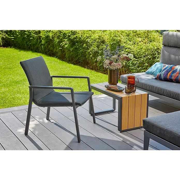 Sunfun Solo Garten-Beistelltisch - L x B x H: 77 x 41,5 x 45 cm, Aluminium, Night-Grey - Ein grauer Gartensessel mit Armlehnen steht auf einer Terrasse, daneben ein Beistelltisch mit Dekoration und einem grauen Sofa.
