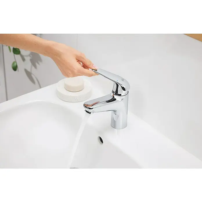 Grohe QuickFix Waschtischarmatur Swift - Chrom, Glänzend, Höhe: 162 mm - Nahaufnahme eines eleganten Waschtischs mit Wasserhahn der Marke Grohe, der von einer Hand bedient wird und Wasser fließen lässt.
