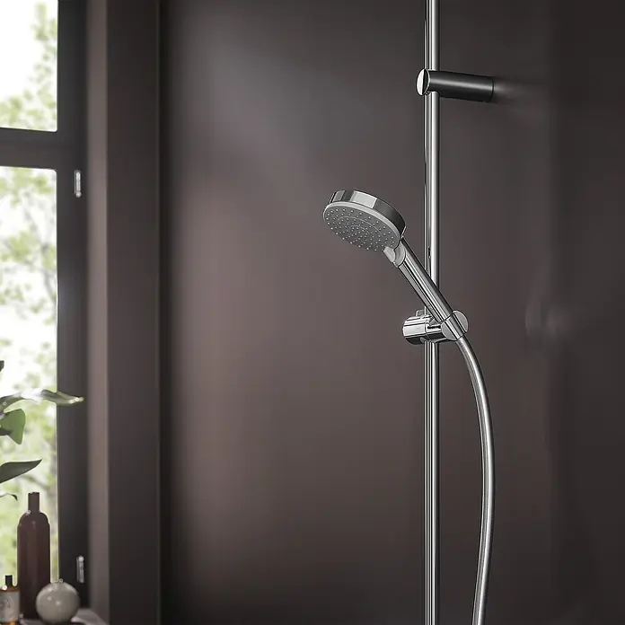 Hansgrohe Vernis Blend Handbrause Vario Eco - Anzahl Funktionen: 2 Stk., Durchmesser: 10 cm, Chrom - Nahaufnahme einer modernen Duschstange aus Chrom mit Handbrause und dunkler Wand im Hintergrund, neben einem Fenster.