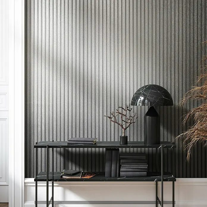 Marburg Travertino Vliestapete Paneel-Optik - Silber, Metalloptik, 10,05 x 0,53 m - Elegantes Interieur mit einer Konsole aus schwarzem Marmor, dekoriert mit Büchern, einer Lampe und einer abstrakten Skulptur vor einer Wand mit vertikalen Linien.