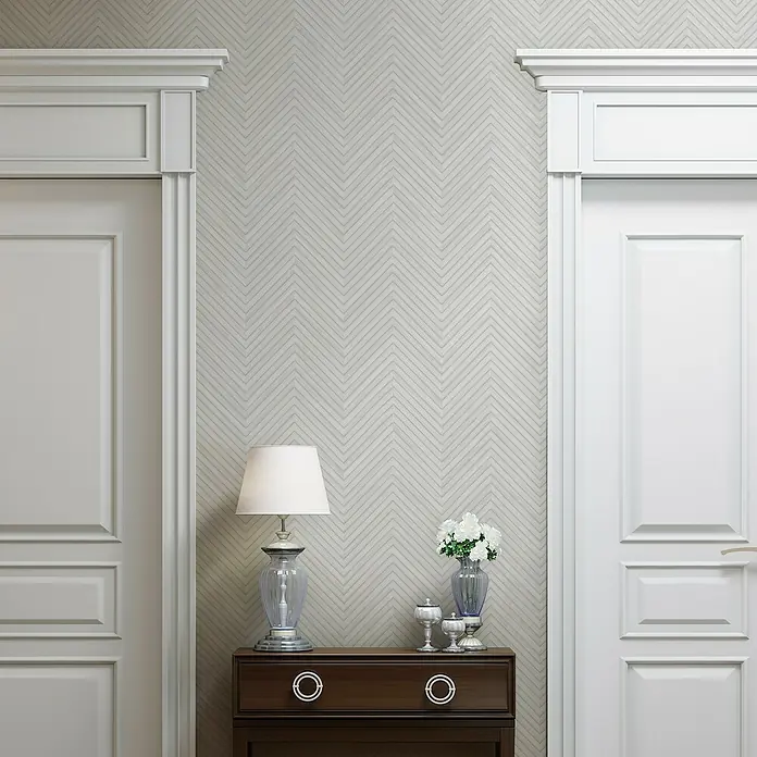Marburg Travertino Vliestapete Fischgrätmuster - Weißgrau, Holzoptik, 10,05 x 0,53 m - Elegante Interieuraufnahme mit einem stilvollen Nachttisch, verziert mit einer Lampe und Blumen, flankiert von weißen Türen.