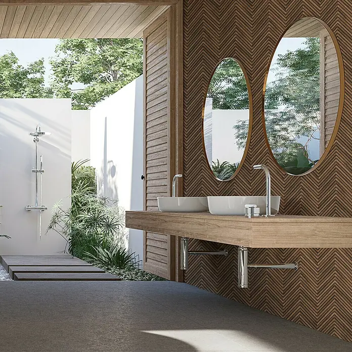 Marburg Travertino Vliestapete Fischgrätmuster - Dunkelbraun, Holzoptik, 10,05 x 0,53 m - Badezimmer mit Holzelementen: Zwei ovale Spiegel über einem Waschtisch aus Holz und ein Außenbereich mit Dusche im Hintergrund.