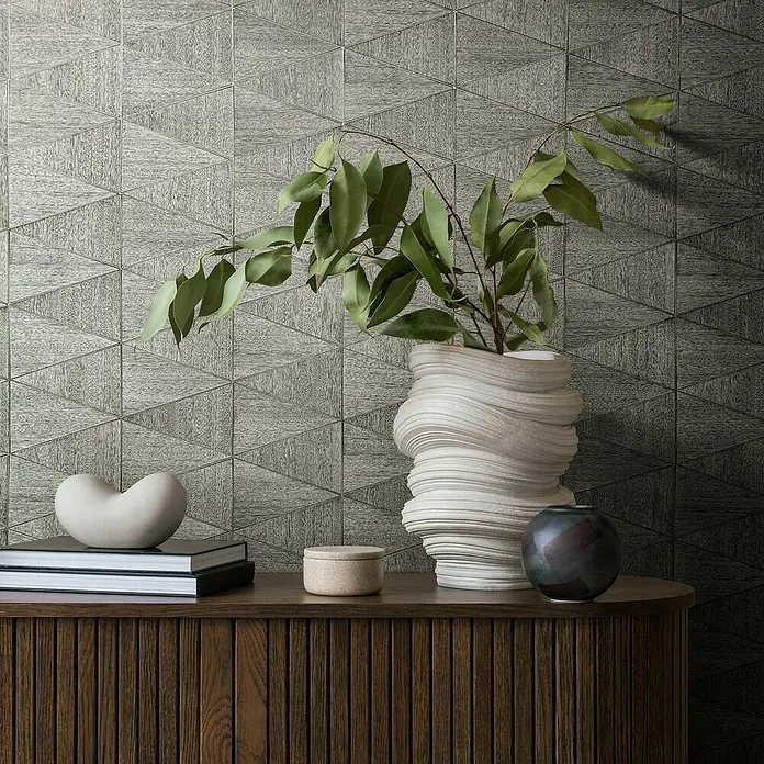Marburg Travertino Papiertapete - Grau, Grafisch, 10,05 x 0,53 m - Dekoratives Arrangement mit einer skulpturalen Vase, Büchern und Keramikobjekten auf einer Holzkommode vor einer geometrischen Wand.