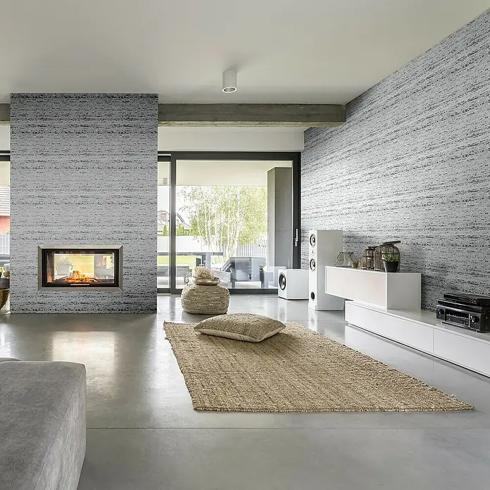 Marburg Travertino Vliestapete Struktur - Hellgrau-Grau, Steinoptik, 10,05 x 0,53 m - Modernes Wohnzimmer mit Teppich, Kamin und Lautsprechern. Das Design ist minimalistisch und stilvoll.
