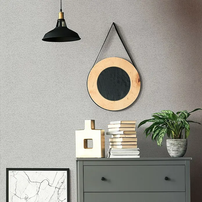 Marburg Travertino Vliestapete Struktur - Grau, Steinoptik, 10,05 x 0,53 m - Nahaufnahme einer stilvollen Kommode mit Dekorationen wie einer Lampe, einem Spiegel, Büchern und einer Pflanze.