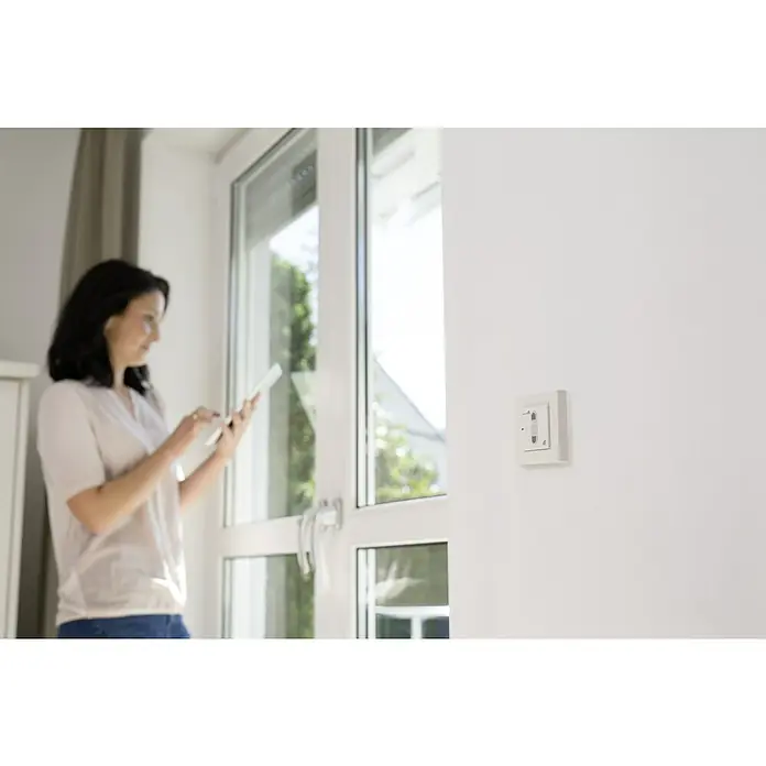 Schellenberg Smart Home Interruptor de recepción por radio - 80 x 80 x 40 mm, Blanco, Montaje enrasado - Un interruptor de luz inteligente en una pared blanca, junto a una mujer con una tableta. Control domótico innovador a la vista.