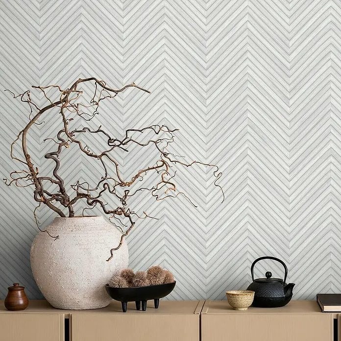 Marburg Travertino Vliestapete Fischgrätmuster - Weißgrau, Holzoptik, 10,05 x 0,53 m - Dekoratives Interieur mit Chevron-Muster-Tapete, Vase mit Ästen, Schale mit Kugeln, Teekanne und Teeschale auf einem Sideboard.