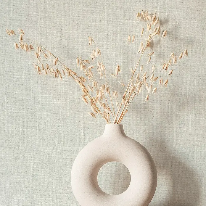 Marburg Travertino Vliestapete - Helles Strukturbeige, Uni, 10,05 x 0,53 m - Nahaufnahme einer stilvollen, beigen Donut-Vase mit zarten, getrockneten Gräsern, die eine schlichte Eleganz für modernes Wohndesign verkörpern.