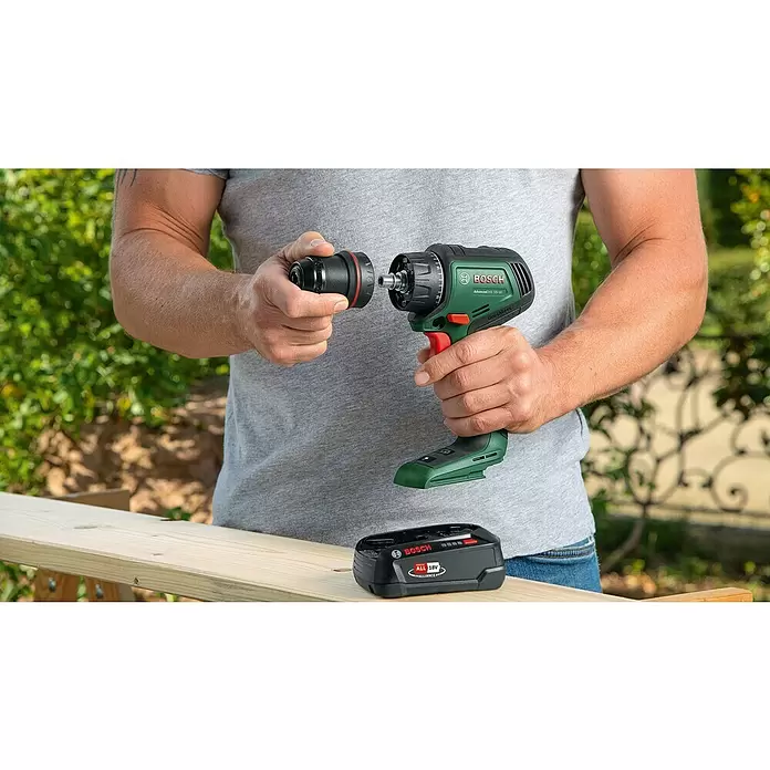 Bosch Power for All 18V Taladro atornillador de batería AdvancedDrill 18V-80 - 18 V, 2 baterías, 2,5 Ah, 78 Nm - Hombre colocando portabrocas negro a taladro verde Bosch AdvancedDrill 18V-80 con detalles rojos. Una batería Bosch 18V descansa en una tabla de madera.