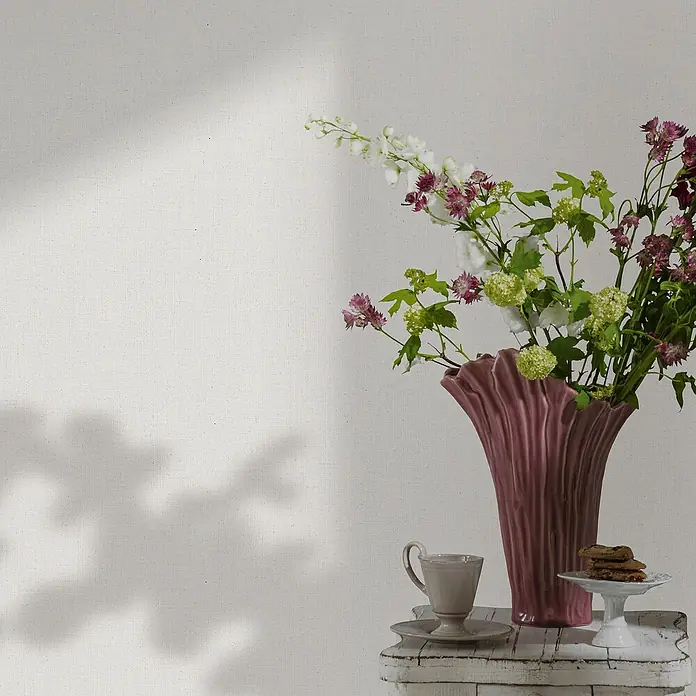 Marburg Travertino Vliestapete - Strukturweiß, Uni, 10,05 x 0,53 m - Ein rosafarbene Vase mit Blumen steht auf einem Tisch, daneben eine Tasse und ein Teller mit Keksen.