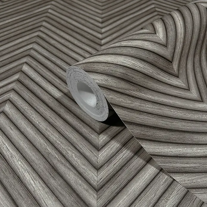 Marburg Travertino Vliestapete Fischgrätmuster - Dunkelgrau, Holzoptik, 10,05 x 0,53 m - Nahaufnahme einer Rolle Chevron-Tapete in Grau. Die Tapete ist mit einem auffälligen, geometrischen Muster versehen, das jedem Raum einen modernen Touch verleiht.