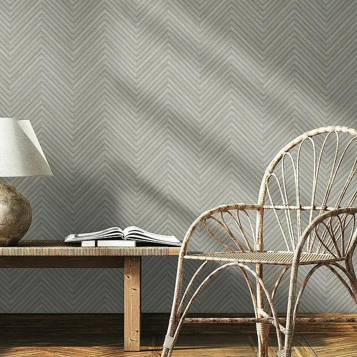 Marburg Travertino Vliestapete Fischgrätmuster - Hellgrau, Holzoptik, 10,05 x 0,53 m - Detailaufnahme eines Raumes mit einem eleganten Rattan-Stuhl vor einer Wand mit Fischgrätenmuster und einer Lampe auf einem Holztisch.
