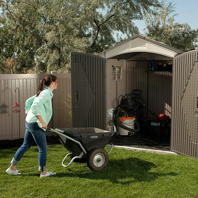 Lifetime Gerätehaus Fox - Außenbreite inkl. Dachüberstand: 215 cm, Außentiefe inkl. Dachüberstand: 210 cm, Kunststoff, Lichtgrau/Anthrazit - Frau schiebt Schubkarre aus einem Lifetime-Gartenschuppen, der Rasenmäher und Werkzeuge enthält. Praktische Aufbewahrung im Freien.