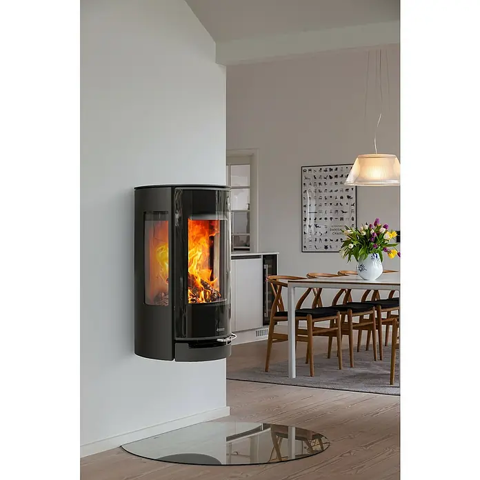 Aduro Kaminofen 22.4 Lux - 5,5 kW, Raumheizvermögen: 150 m³, Material Verkleidung: Stahl, Schwarz - Ein moderner, an der Wand montierter Kaminofen mit Glasfront, der das brennende Holz zeigt, steht in einem stilvollen Esszimmer.