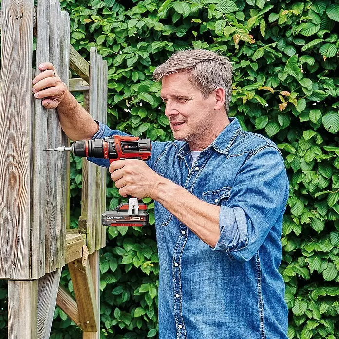 Einhell PXC 18V Taladro atornillador de batería TE-CD 18/45 3X-Li - 18 V, 1 batería, 2 Ah - Hombre usa taladro atornillador Einhell rojo y negro en una estructura de madera exterior.