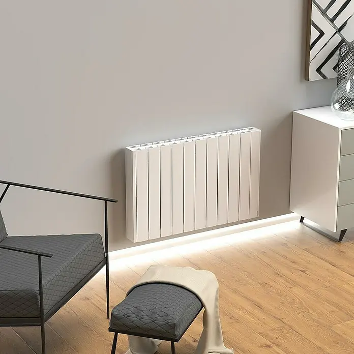 Rointe Emisor térmico Belize WiFi - 1.100 W, Blanco, 9,5 x 90 x 57,5 cm, Fluido - Un radiador moderno blanco está montado en una pared gris, iluminado desde abajo, junto a un sillón gris y una cómoda.