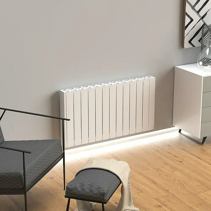 Rointe Emisor térmico Belize WiFi - 1.200 W, Blanco, 9,5 x 106 x 57,5 cm, Fluido - Un radiador blanco moderno con aletas elegantes montado en una pared gris, acentuado por un cálido resplandor debajo.