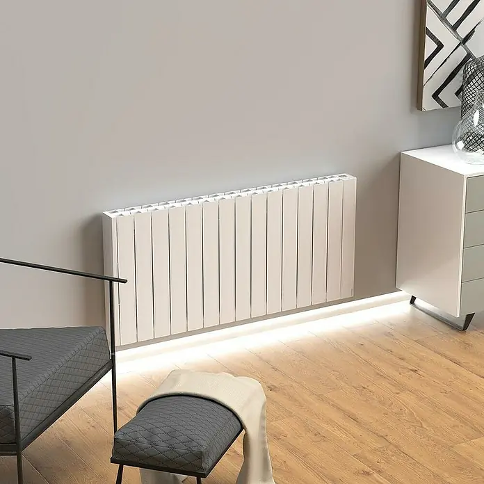 Rointe Emisor térmico Belize WiFi - 1.540 W, Blanco, 9,5 x 122 x 57,5 cm, Fluido - Primer plano de un radiador blanco contra una pared gris, con una silla y un armario al fondo.