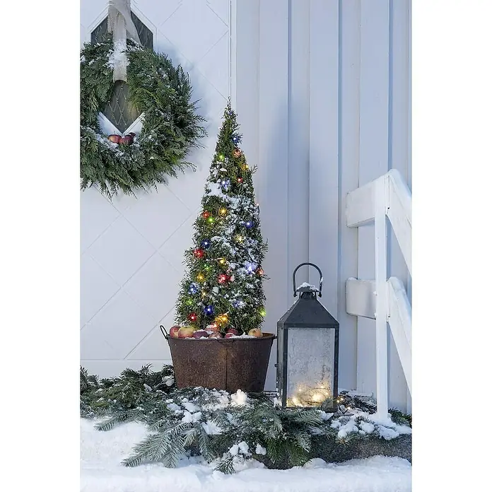 Konstsmide LED-Lichterkette - Außen, Lichtfarbe: Mehrfarbig, Anzahl Leuchtmittel: 80 Stk., Netzbetrieben, 12,64 m - Winterliche Außendekoration mit beleuchtetem Baum im Kübel, Kranz an der Tür und einer Laterne, alles bedeckt mit leichtem Schnee.