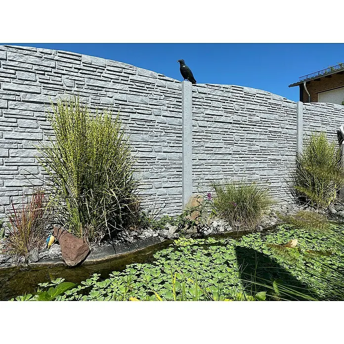 Beckers Betonzaun Abschlussplatte Standard Montanabogen - 200 x 45 cm, Beton, Betongrau - Blick auf einen Gartenteich mit Seerosen und einer grauen Steinmauer im Hintergrund, auf der ein Vogel sitzt.