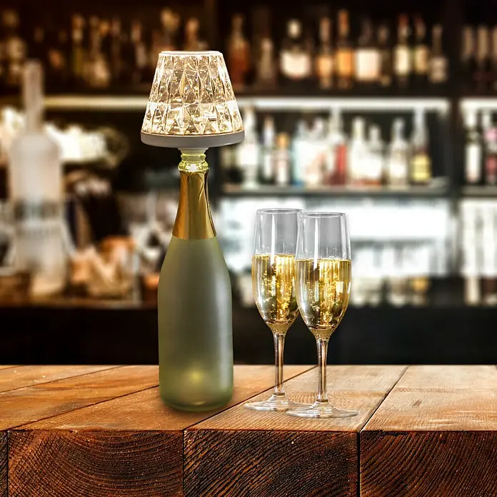 REV LED-Flaschenleuchte Lamprusco Cristal - 2 W, Weiß, Warmweiß - Nahaufnahme einer Champagnerflasche mit Lampe auf einem Holztisch neben zwei Gläsern mit Champagner, im Hintergrund eine Bar.