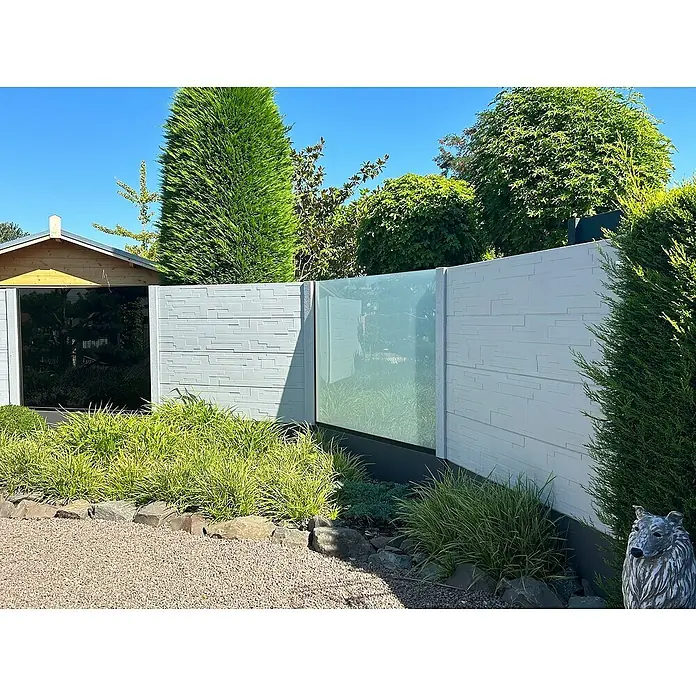 Beckers Betonzaun Zaunelement Klassik Rimini Motiv - 184 x 26 cm, Beton, Betongrau - Bild einer modernen, grauen Gartenzaun-Installation mit Glaselementen, umgeben von üppigem Grün und einem dekorativen Steingarten.