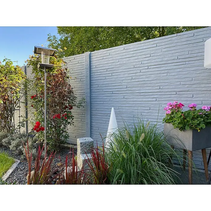 Beckers Betonzaun Zaunelement Standard Toscana Motiv - 200 x 38,5 cm, Beton, Betongrau - Eine graue Betonzaunplatte mit horizontalem Ziegeldesign schmückt einen Garten mit Blumen und dekorativen Elementen.