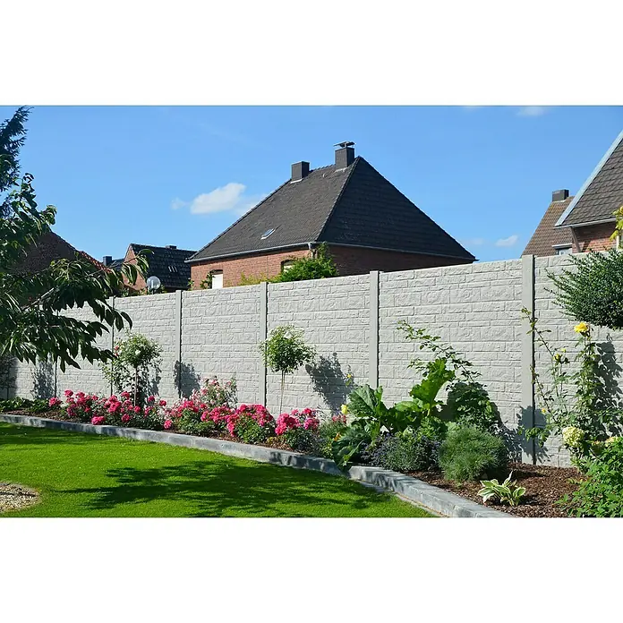 Beckers Betonzaun Zaunelement Standard Fels Motiv - 200 x 38,5 cm, Beton, Betongrau - Graue Betonzaunpaneele mit Ziegelmuster grenzen einen grünen Garten mit Rosenbeeten und Sträuchern ab, Häuser im Hintergrund.