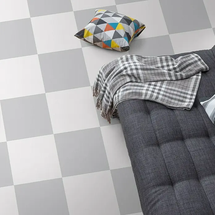 Feinsteinzeugfliese Barcelona Gris - 24,8 x 24,8 cm, Grau, Matt - Bild eines karierten Fussbodens mit einem Sofa, einem Kissen und einer Decke. Das Sofa ist grau und hat eine Knopfheftung.