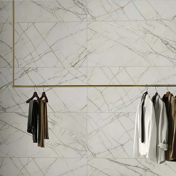 Feinsteinzeugfliese Statuario Shining - 60 x 120 cm, Weiß/Grau, Matt - Eine Marmorwand mit einer goldenen Kleiderstange, an der Hemden und Hosen hängen, die eine stilvolle Präsentation der angebotenen Produkte bietet.